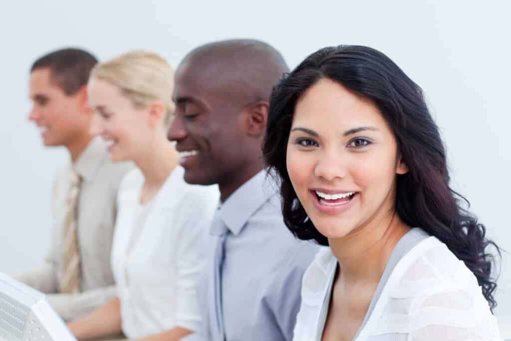 Diversity-Initiativen: Junge Frau lächelt in die Kamera, während mehrere Kolleginnen und Kollegen am Computer arbeiten.
