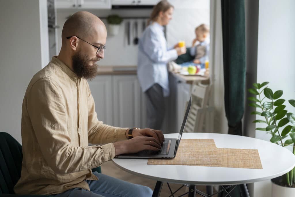Mann arbeitet im Homeoffice an einem Laptop, im Hintergrund seine Familie in der Küche